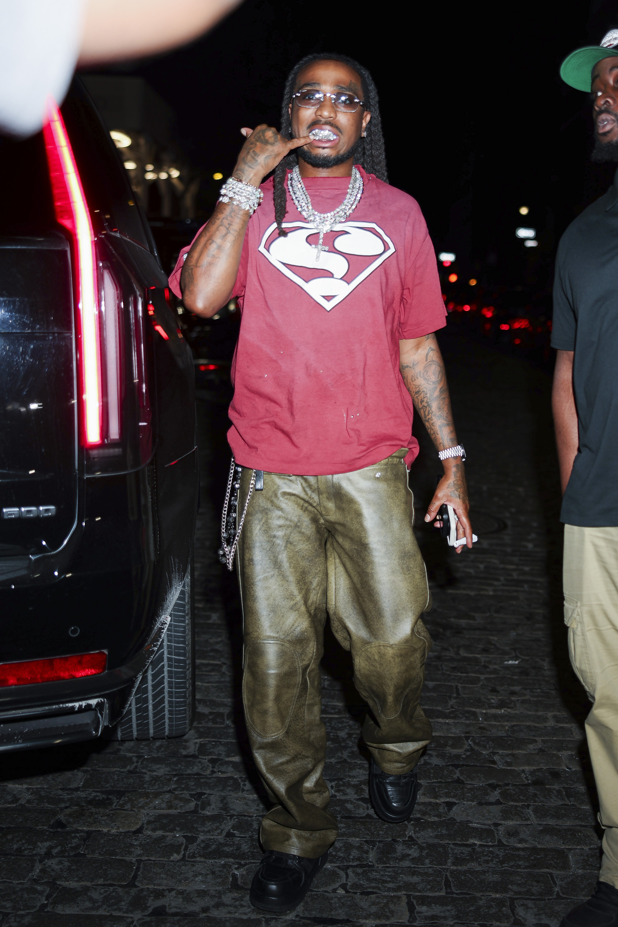 Man in a red Superman shirt and green pants, wearing sunglasses and jewelry, stands next to a black car at night.
