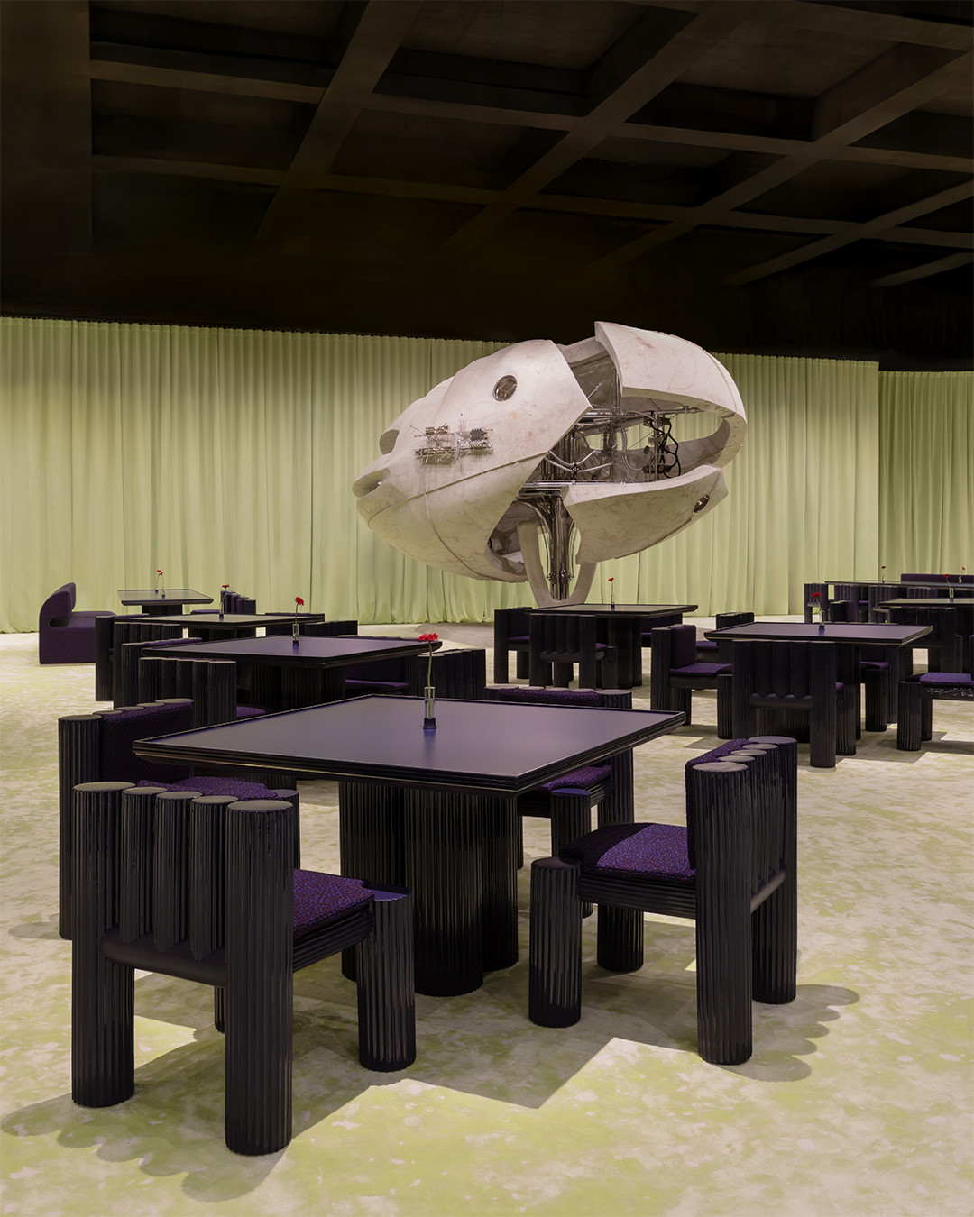 A modern dining space with black tables and chairs, featuring a futuristic, spherical sculpture suspended above. Green curtains in the background.