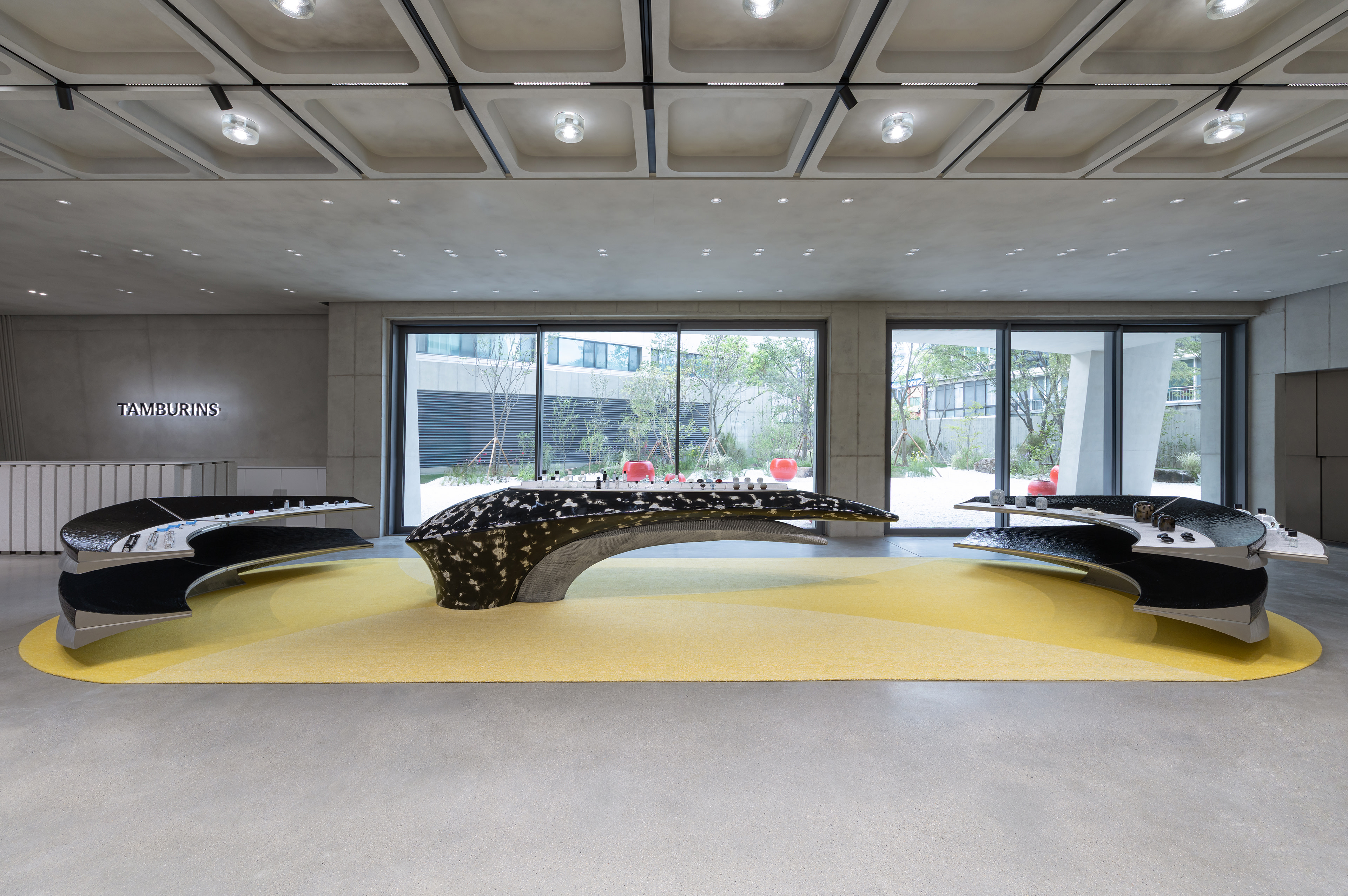 Modern showroom interior with sleek, curved display tables on a yellow carpet. Large windows reveal a garden outside. Branding reads 