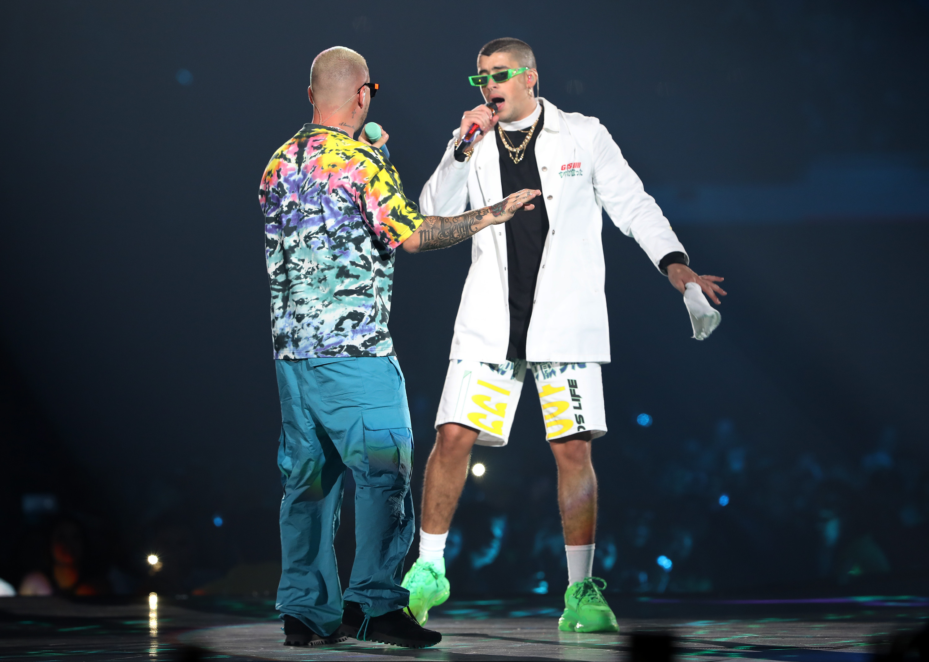 Two performers on stage, one in a colorful shirt and the other in a white coat and green shoes, during a live concert.