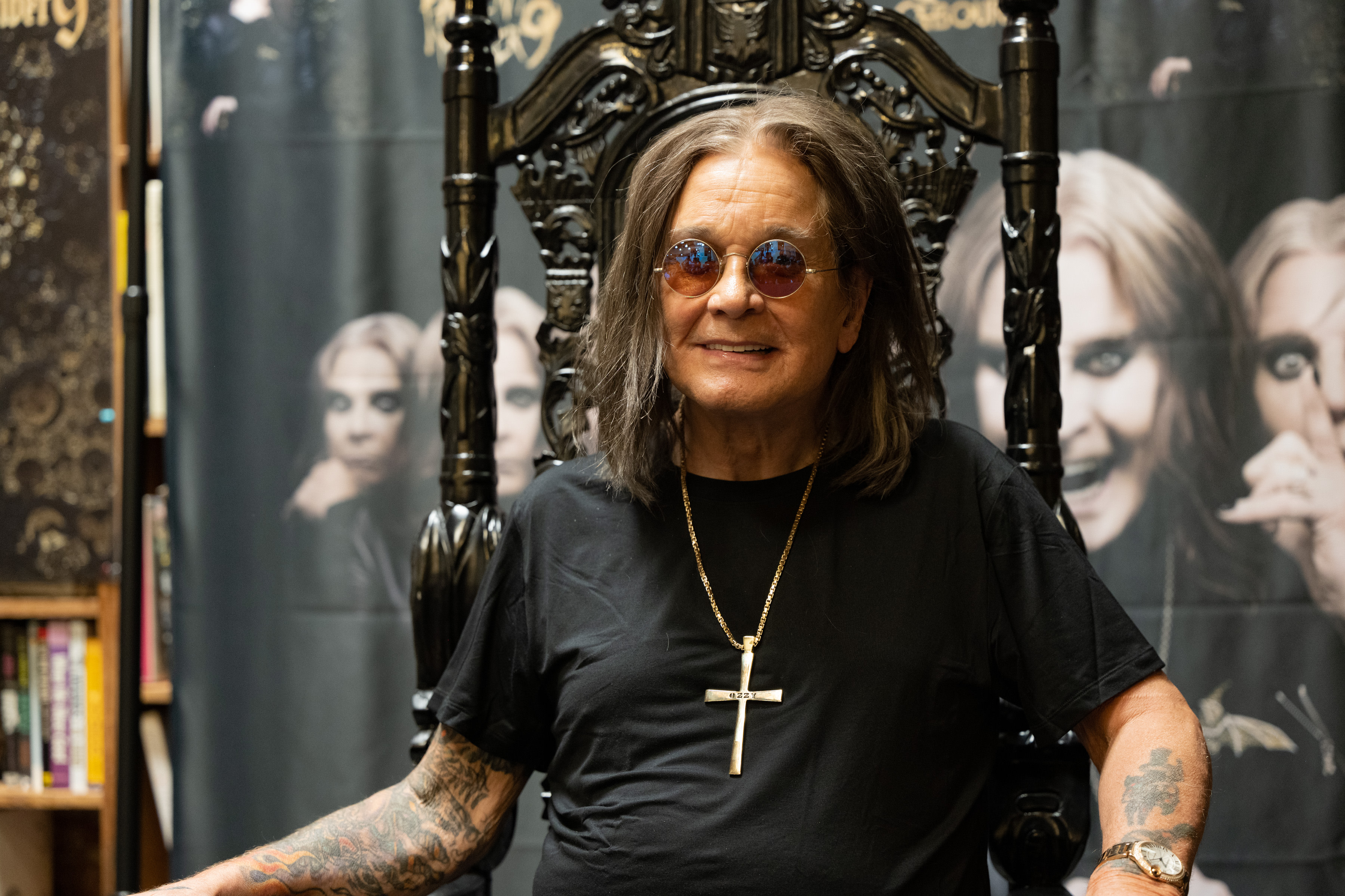 Ozzy Osbourne sitting on a throne-like chair, wearing sunglasses and a cross necklace, with a backdrop featuring his images.