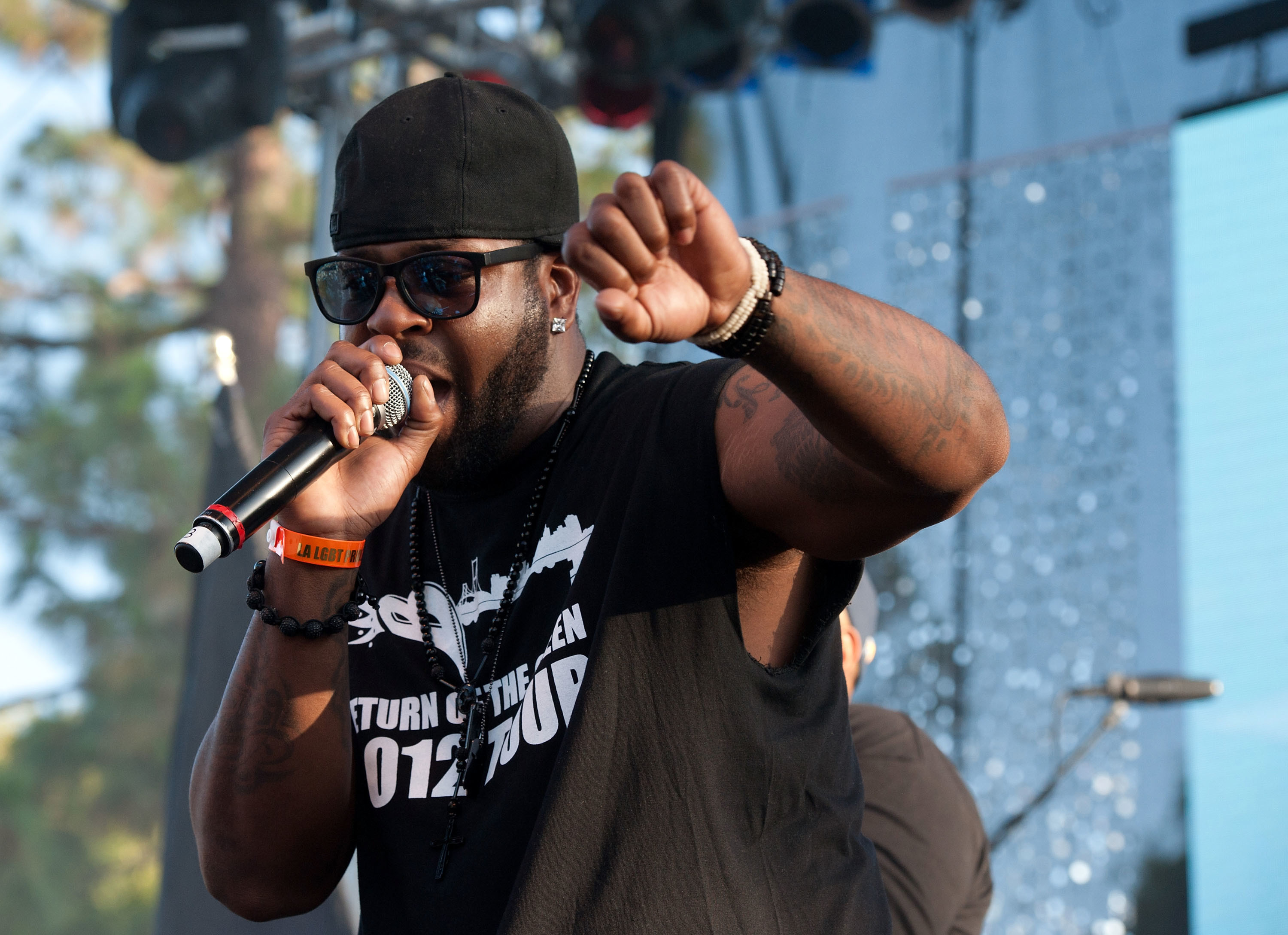 A man wearing sunglasses and a black cap performs on stage, holding a microphone and raising his fist, with blurred stage lights behind him.