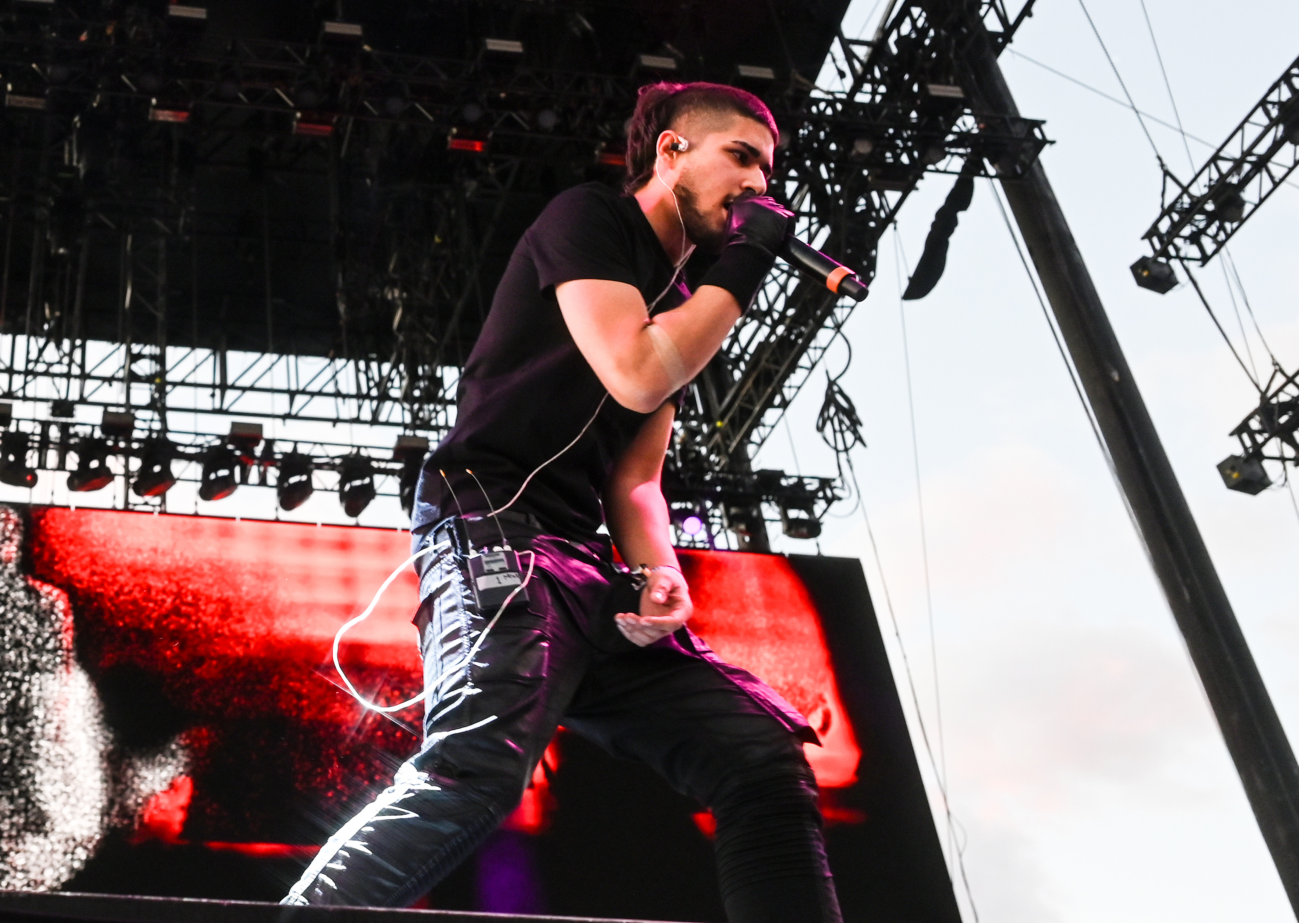 fakemink on stage holding a microphone, wearing a black outfit and gloves, with a large screen and stage lights in the background.