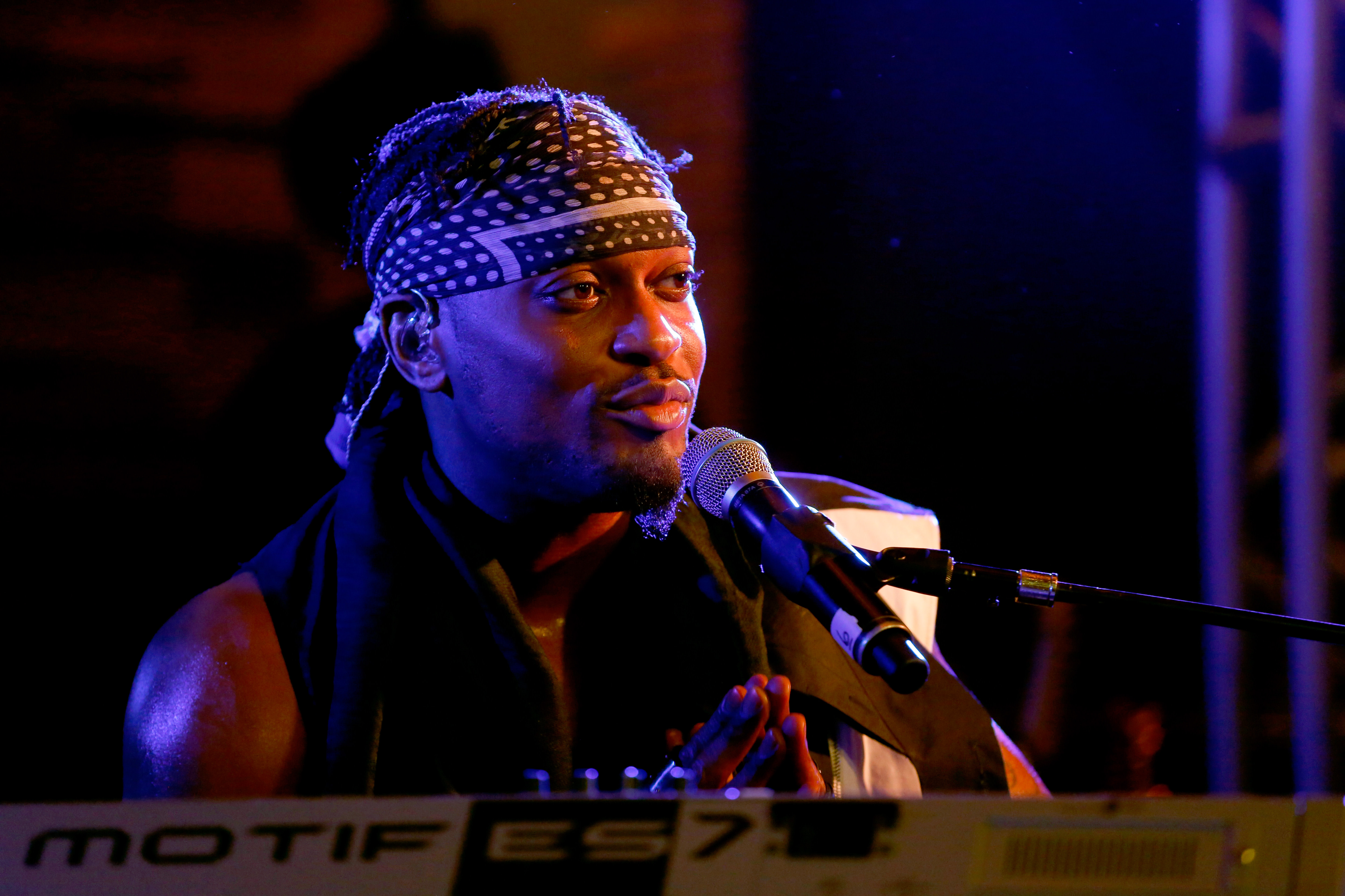 A musician wearing a bandana and scarf performs on stage, playing a keyboard and singing into a microphone under blue lighting.