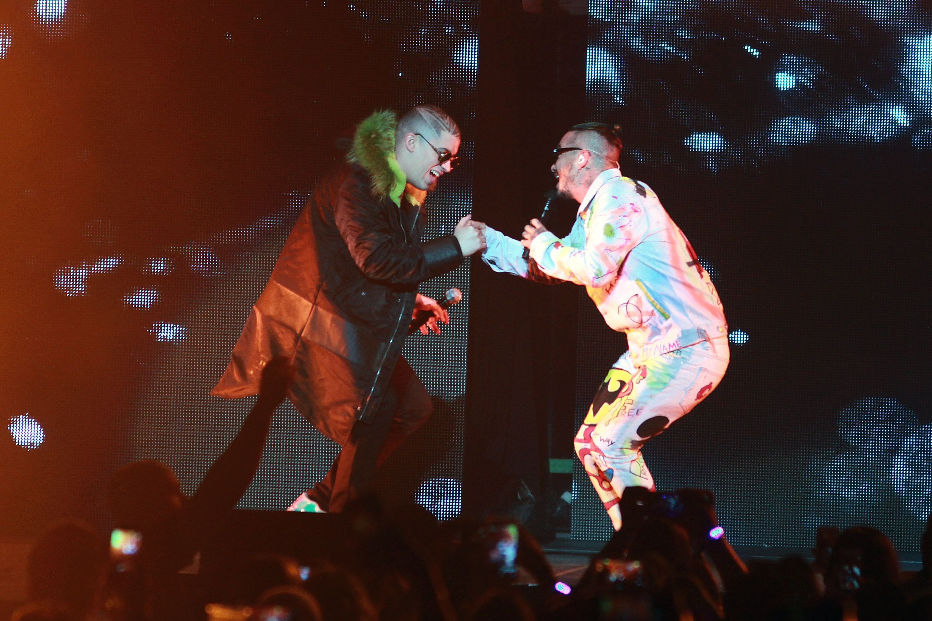 Two performers on stage, one in a black coat with green fur, the other in a colorful suit, interacting energetically under bright lights.
