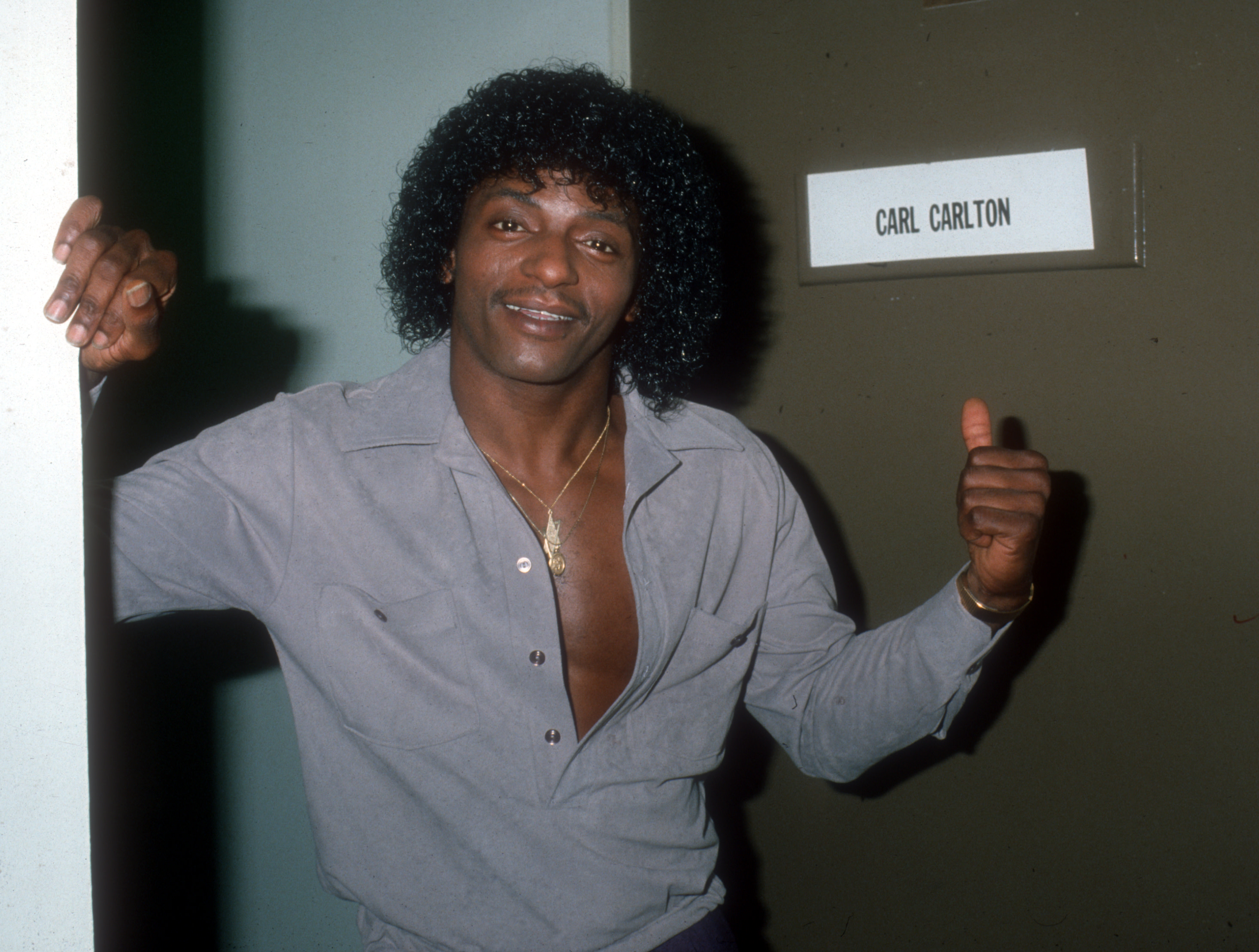Carl Carlton smiling, giving a thumbs up, standing by a door with his name on it, wearing a gray shirt and gold jewelry.