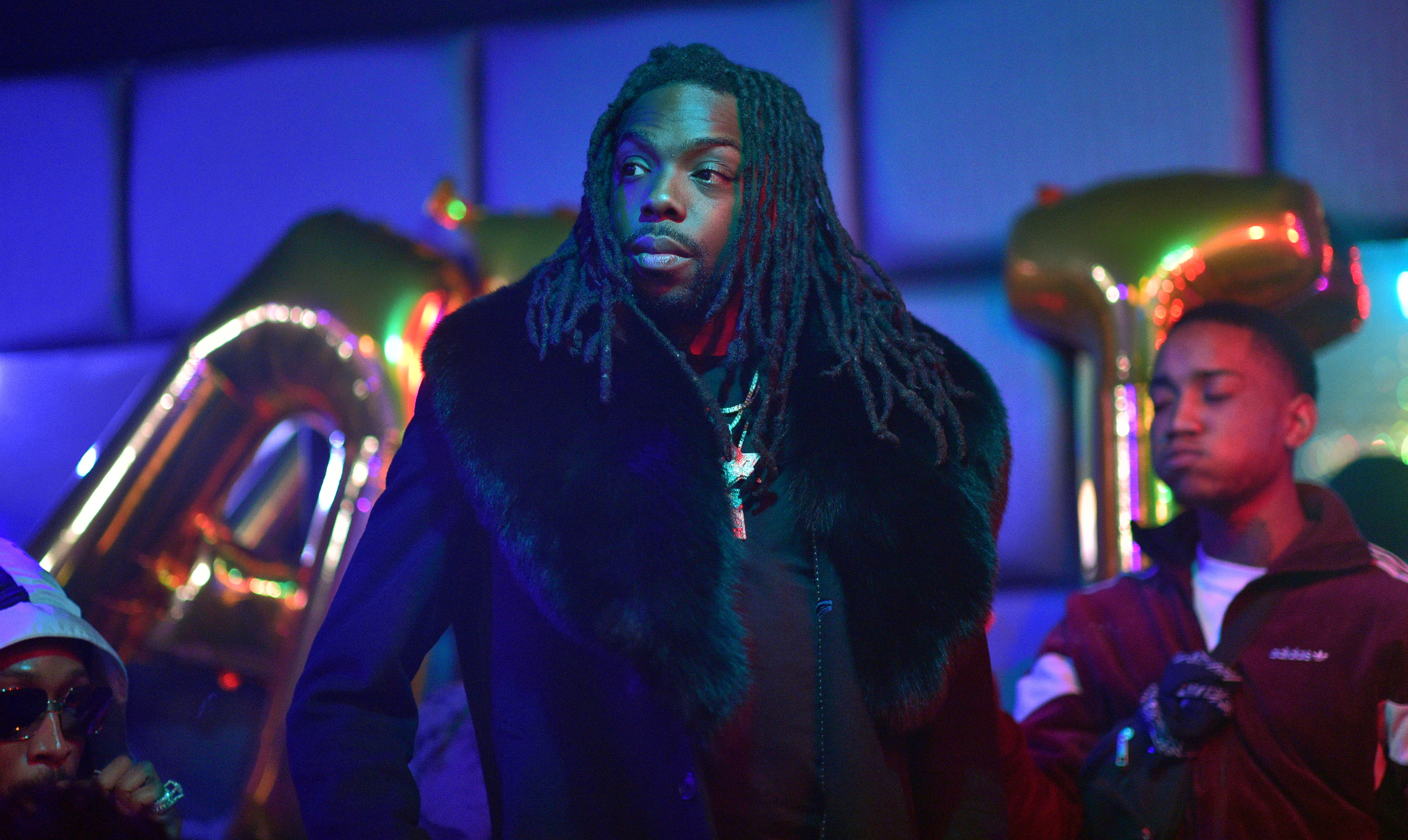 A man with long dreadlocks and a fur coat stands in a dimly lit room with colorful lighting and balloons in the background.