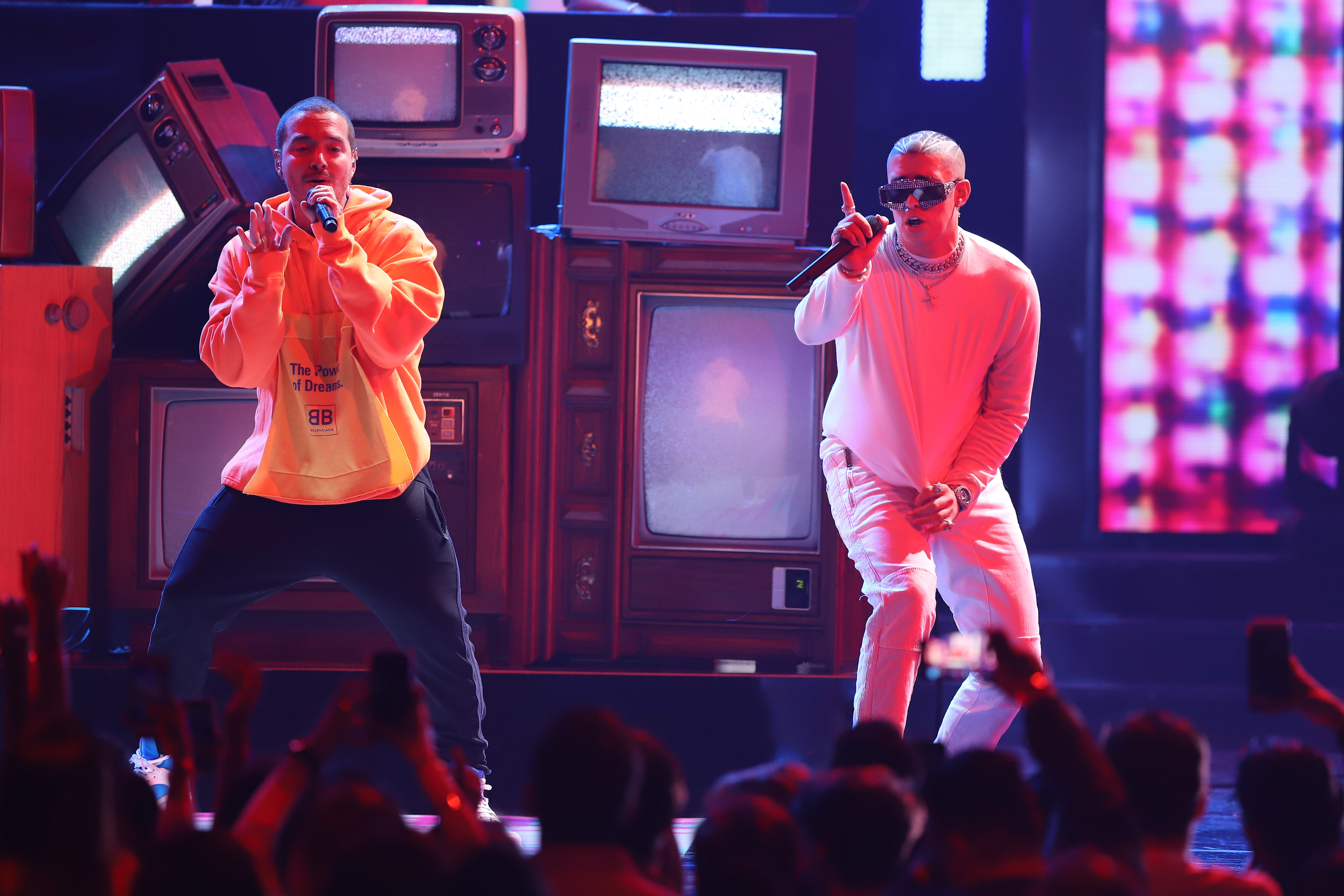 Two performers on stage with vintage TVs in the background, one in a yellow hoodie and the other in white, singing to a crowd.