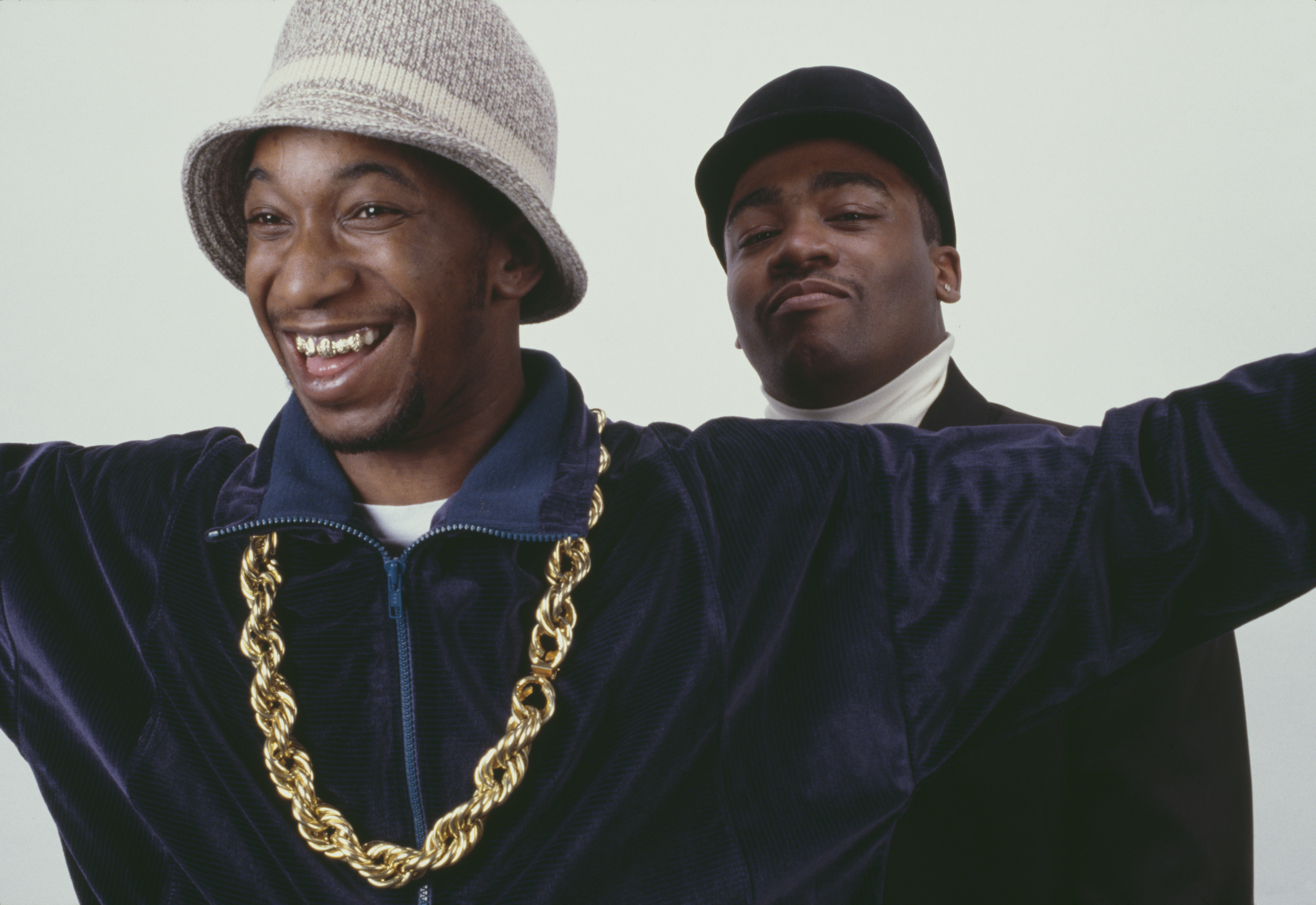 Two men smiling; one wears a bucket hat and gold chain, the other in a black cap and turtleneck. Retro hip-hop style.