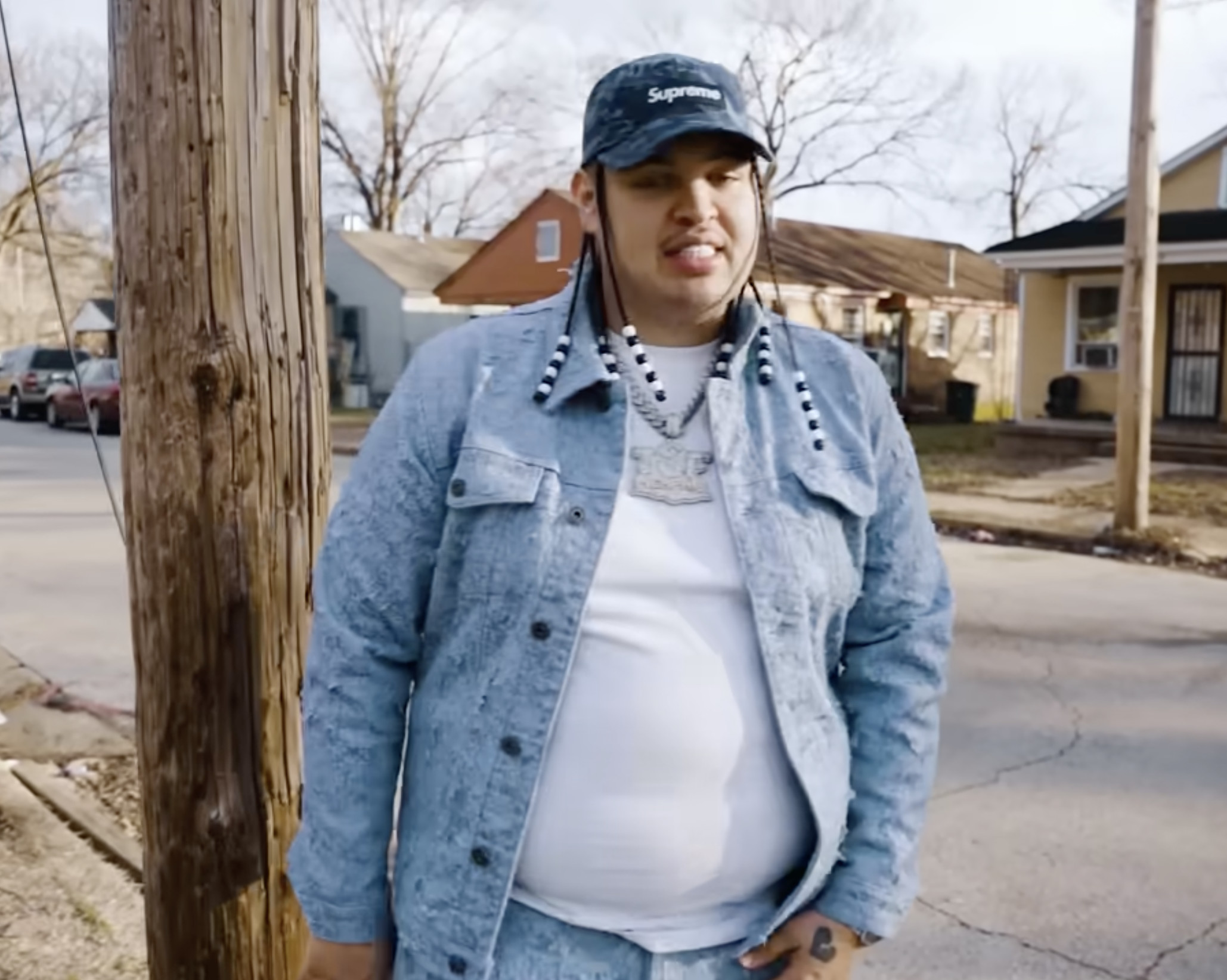 A person wearing a denim jacket, white shirt, and a Supreme cap stands by a utility pole on a residential street.