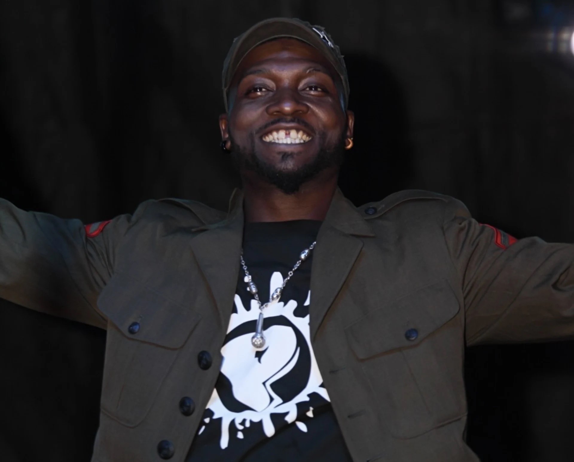 A smiling man in a dark jacket and cap, wearing a necklace and graphic t-shirt, arms raised in a celebratory gesture.