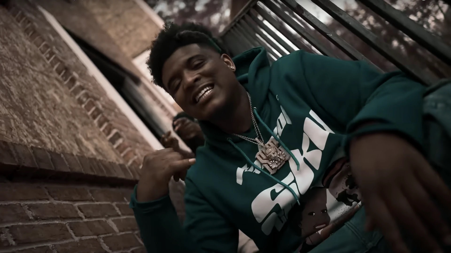 A person in a teal hoodie with a large pendant necklace, sitting on outdoor steps, smiling and gesturing with their hand.