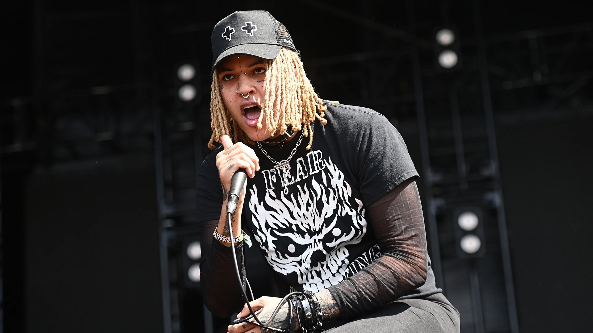 A performer with blonde dreadlocks and a black cap sings passionately on stage, wearing a black shirt with a white graphic.