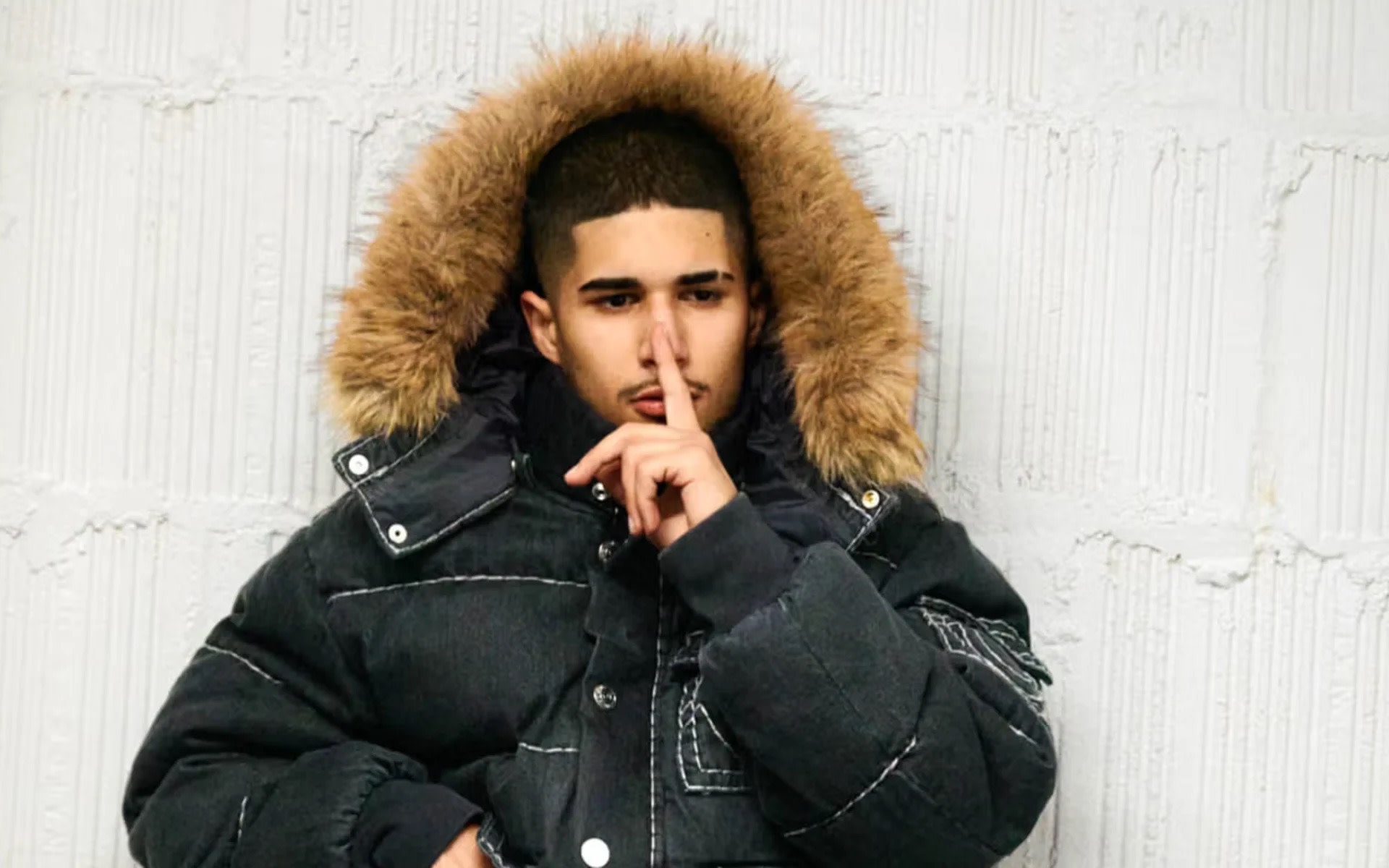 uk rapper fakemink in a thick jacket with a fur-lined hood, holding a finger to their lips against a textured wall background.